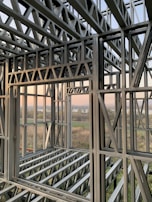 A complex structure of metal beams and girders forming a framework. The geometric design with interlocking beams suggests an in-progress construction, with a scenic view visible through the open spaces between the metal framework. The landscape outside includes fields and a water body under a clear sky.