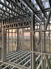 A complex structure of metal beams and girders forming a framework. The geometric design with interlocking beams suggests an in-progress construction, with a scenic view visible through the open spaces between the metal framework. The landscape outside includes fields and a water body under a clear sky.