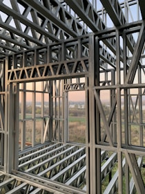 A complex structure of metal beams and girders forming a framework. The geometric design with interlocking beams suggests an in-progress construction, with a scenic view visible through the open spaces between the metal framework. The landscape outside includes fields and a water body under a clear sky.