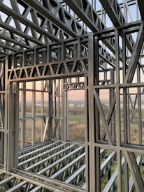 A complex structure of metal beams and girders forming a framework. The geometric design with interlocking beams suggests an in-progress construction, with a scenic view visible through the open spaces between the metal framework. The landscape outside includes fields and a water body under a clear sky.
