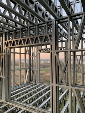 A complex structure of metal beams and girders forming a framework. The geometric design with interlocking beams suggests an in-progress construction, with a scenic view visible through the open spaces between the metal framework. The landscape outside includes fields and a water body under a clear sky.