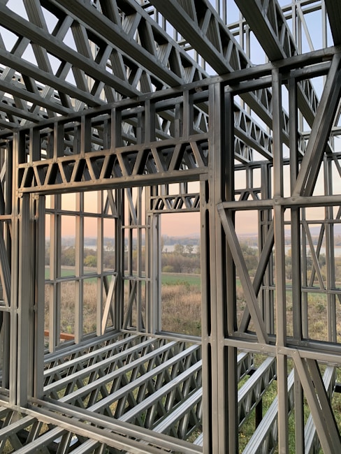 A complex structure of metal beams and girders forming a framework. The geometric design with interlocking beams suggests an in-progress construction, with a scenic view visible through the open spaces between the metal framework. The landscape outside includes fields and a water body under a clear sky.