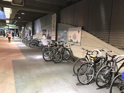 A row of bicycles is parked neatly against the wall in a well-lit indoor area, which appears to be a train or bus station. The walls are lined with advertisements, and there are a few people walking in the background with a visible sign leading to another area.
