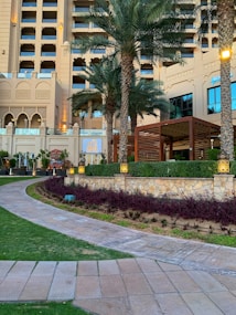 A luxurious hotel exterior featuring a multi-story classical architectural design with large windows and ornate details. Tall palm trees are planted around a beautifully landscaped garden with pathways. A restaurant sign named 'Miss India' is visible, and ambient lighting enhances the elegant setting.