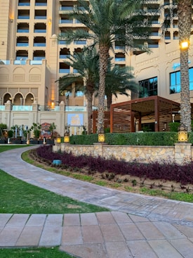 A luxurious hotel exterior featuring a multi-story classical architectural design with large windows and ornate details. Tall palm trees are planted around a beautifully landscaped garden with pathways. A restaurant sign named 'Miss India' is visible, and ambient lighting enhances the elegant setting.