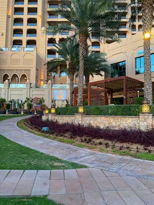 A luxurious hotel exterior featuring a multi-story classical architectural design with large windows and ornate details. Tall palm trees are planted around a beautifully landscaped garden with pathways. A restaurant sign named 'Miss India' is visible, and ambient lighting enhances the elegant setting.