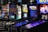 Pinball machines with colorful displays featuring themes like Jurassic Park, Willy Wonka, and Stranger Things are situated in an arcade-like setting. Bright lights and neon colors illuminate the machines, creating a dynamic and engaging atmosphere. In the background, vending machines and a variety of snacks and beverages are visible.