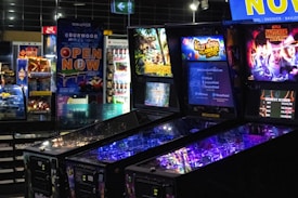 Pinball machines with colorful displays featuring themes like Jurassic Park, Willy Wonka, and Stranger Things are situated in an arcade-like setting. Bright lights and neon colors illuminate the machines, creating a dynamic and engaging atmosphere. In the background, vending machines and a variety of snacks and beverages are visible.