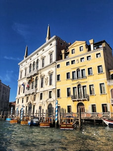 A Venetian canal scene featuring a historic building with ornate architecture and tall spires. The building has yellow and white facades with numerous windows. In the foreground, several wooden boats labeled 'Regione Veneto' are docked next to striped blue and white mooring posts. The water reflects the clear blue sky.