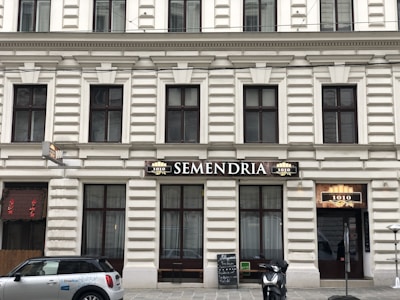 A building facade with classic architectural features, including rectangular windows and decorative moldings. The central focus is a sign with the word 'SEMENDRIA' above a doorway. A scooter is parked on the sidewalk, and a car with 'DriveNow' branding is visible. A blackboard menu is placed outside, beside a door with the number 1010.