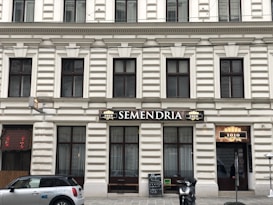 A building facade with classic architectural features, including rectangular windows and decorative moldings. The central focus is a sign with the word 'SEMENDRIA' above a doorway. A scooter is parked on the sidewalk, and a car with 'DriveNow' branding is visible. A blackboard menu is placed outside, beside a door with the number 1010.