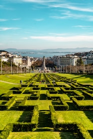 Geometric hedges form a pattern on a spacious public park with people walking and relaxing. In the background, urban buildings and a distant view of a body of water are visible along with rolling hills under a clear blue sky.