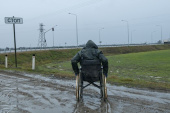A person in a wheelchair, covered in a raincoat, is facing a wet and muddy path near a road. The scene is overcast with a cloudy sky, and there is a 'stop' sign written in Cyrillic next to the person. In the background, there is a traffic light and power lines along with a highway bordered by grass.