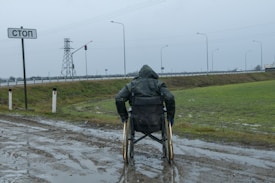 A person in a wheelchair, covered in a raincoat, is facing a wet and muddy path near a road. The scene is overcast with a cloudy sky, and there is a 'stop' sign written in Cyrillic next to the person. In the background, there is a traffic light and power lines along with a highway bordered by grass.