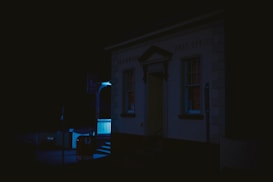 A dimly lit post office building at night, with a faint blue glow illuminating the entrance and a sign nearby. The scene is predominantly dark, highlighting the structural features of the building.