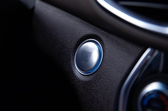 A close-up view of a car's engine start/stop button located on a sleek, dark dashboard. The button is metallic and circular, surrounded by a textured surface.