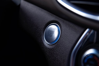 A close-up view of a car's engine start/stop button located on a sleek, dark dashboard. The button is metallic and circular, surrounded by a textured surface.
