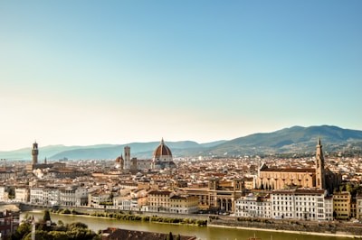 A panoramic view of a historic Italian city with a river in the foreground. The architecture features red-tiled roofs and prominent landmarks like a large domed cathedral and a tall clock tower. Rolling hills can be seen in the background under a clear blue sky.
