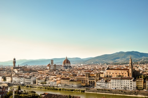 A panoramic view of a historic Italian city with a river in the foreground. The architecture features red-tiled roofs and prominent landmarks like a large domed cathedral and a tall clock tower. Rolling hills can be seen in the background under a clear blue sky.