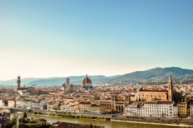 A panoramic view of a historic Italian city with a river in the foreground. The architecture features red-tiled roofs and prominent landmarks like a large domed cathedral and a tall clock tower. Rolling hills can be seen in the background under a clear blue sky.