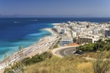 A scenic coastal city with white buildings densely packed near the shoreline. A winding road curves down towards the town, offering a view of the clear blue sea and sandy beach lined with umbrellas. In the background, mountains can be seen across the water under a clear sky.