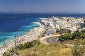 A scenic coastal city with white buildings densely packed near the shoreline. A winding road curves down towards the town, offering a view of the clear blue sea and sandy beach lined with umbrellas. In the background, mountains can be seen across the water under a clear sky.