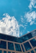A modern building with reflective glass windows against a bright blue sky dotted with white clouds.