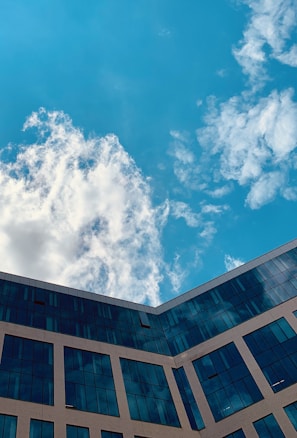 A modern building with reflective glass windows against a bright blue sky dotted with white clouds.