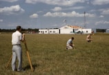 Three men are working on a green field; one is operating a theodolite on a tripod, while the other two are kneeling on the grass, marking or measuring points. In the background, a small airplane is parked in front of a hangar, which has bold numbers painted on its roof. The sky is partly cloudy, and there is a tall communications or weather tower near the hangar.
