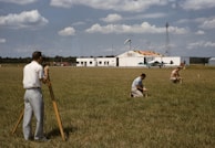 Three men are working on a green field; one is operating a theodolite on a tripod, while the other two are kneeling on the grass, marking or measuring points. In the background, a small airplane is parked in front of a hangar, which has bold numbers painted on its roof. The sky is partly cloudy, and there is a tall communications or weather tower near the hangar.