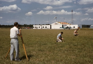 Three men are working on a green field; one is operating a theodolite on a tripod, while the other two are kneeling on the grass, marking or measuring points. In the background, a small airplane is parked in front of a hangar, which has bold numbers painted on its roof. The sky is partly cloudy, and there is a tall communications or weather tower near the hangar.