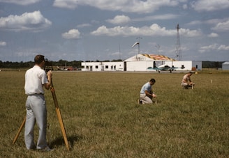 Three men are working on a green field; one is operating a theodolite on a tripod, while the other two are kneeling on the grass, marking or measuring points. In the background, a small airplane is parked in front of a hangar, which has bold numbers painted on its roof. The sky is partly cloudy, and there is a tall communications or weather tower near the hangar.