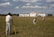 Three men are working on a green field; one is operating a theodolite on a tripod, while the other two are kneeling on the grass, marking or measuring points. In the background, a small airplane is parked in front of a hangar, which has bold numbers painted on its roof. The sky is partly cloudy, and there is a tall communications or weather tower near the hangar.