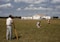 Three men are working on a green field; one is operating a theodolite on a tripod, while the other two are kneeling on the grass, marking or measuring points. In the background, a small airplane is parked in front of a hangar, which has bold numbers painted on its roof. The sky is partly cloudy, and there is a tall communications or weather tower near the hangar.