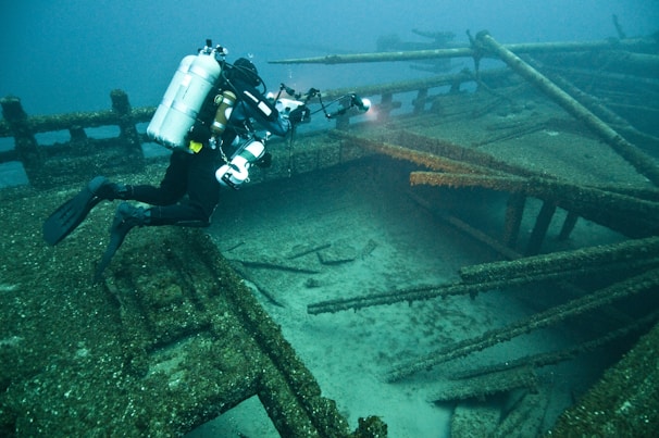 A scuba diver is exploring the wreckage of a sunken ship underwater. The diver, equipped with a full set of scuba gear including a large oxygen tank, is shining a flashlight into the depths of the ship. The surrounding area is murky, and the ship's structure is covered in algae and marine growth.