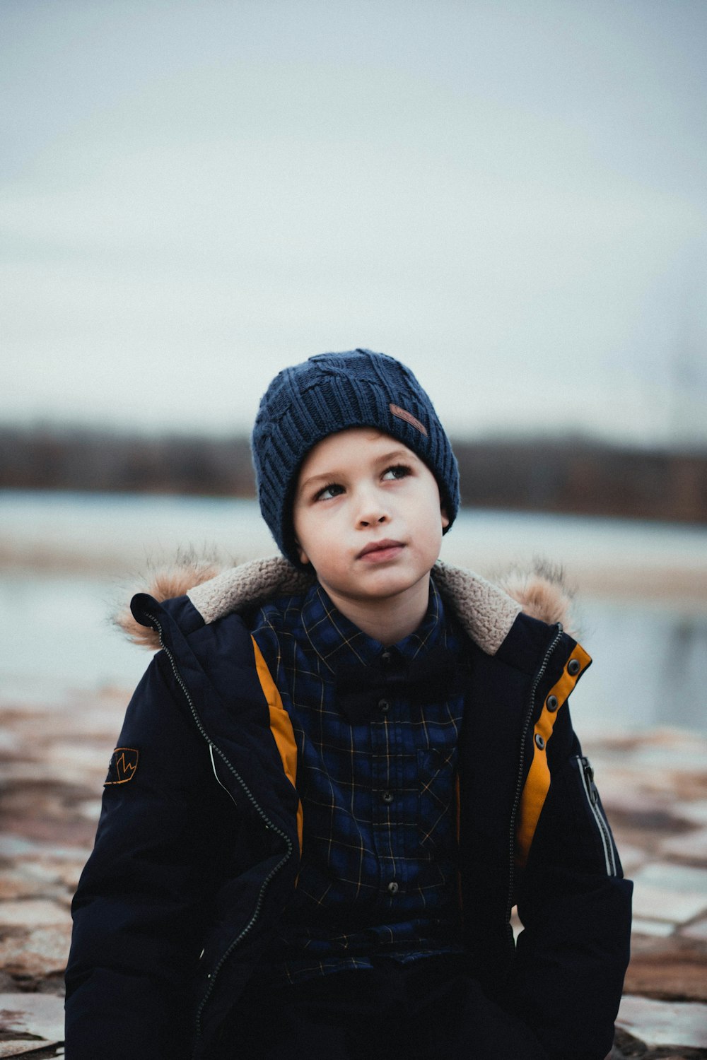 boy in black and yellow jacket and knit cap standing near body of water during daytime
