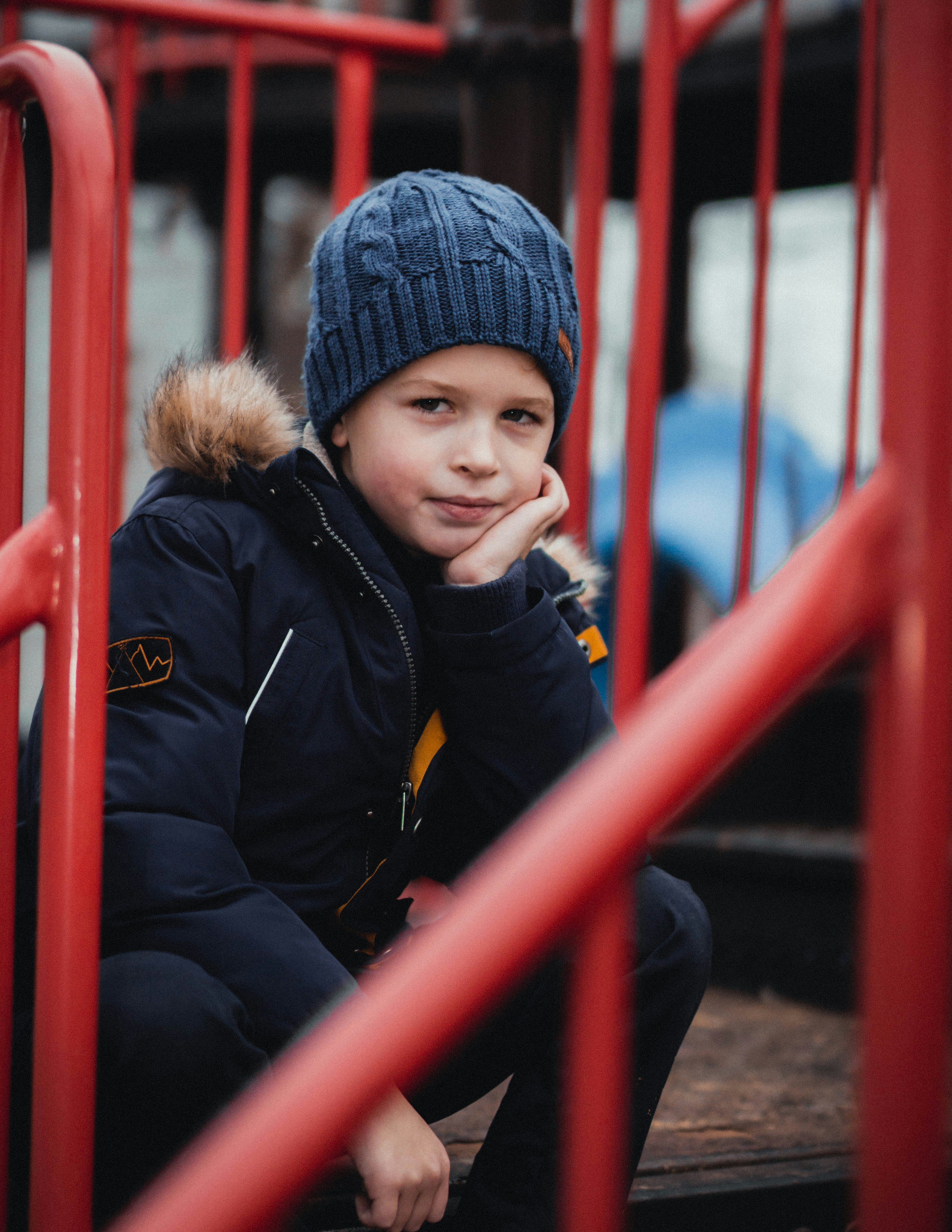 Niño con chaqueta negra y gorra de punto azul de pie junto a la valla de metal rojo durante el día