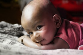 A baby with a gentle smile, resting on its stomach with its head turned towards the camera. The baby wears a sleeveless garment, and a soft light highlights its smooth skin. The background is blurred, emphasizing the baby's expressive eyes and serene expression.