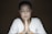 A person with short dark hair and a serene expression is seated with eyes closed and hands clasped in a prayer-like gesture. The lighting is soft, creating a calm and introspective atmosphere, and the background is dark brown, which draws attention to the individual in the center.