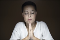 A person with short dark hair and a serene expression is seated with eyes closed and hands clasped in a prayer-like gesture. The lighting is soft, creating a calm and introspective atmosphere, and the background is dark brown, which draws attention to the individual in the center.