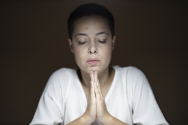 A person with short dark hair and a serene expression is seated with eyes closed and hands clasped in a prayer-like gesture. The lighting is soft, creating a calm and introspective atmosphere, and the background is dark brown, which draws attention to the individual in the center.