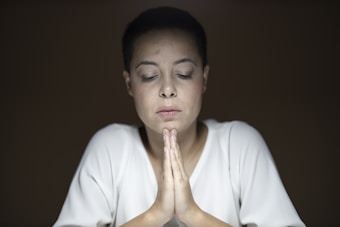 A person with short dark hair and a serene expression is seated with eyes closed and hands clasped in a prayer-like gesture. The lighting is soft, creating a calm and introspective atmosphere, and the background is dark brown, which draws attention to the individual in the center.