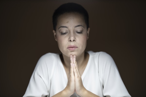 A person with short dark hair and a serene expression is seated with eyes closed and hands clasped in a prayer-like gesture. The lighting is soft, creating a calm and introspective atmosphere, and the background is dark brown, which draws attention to the individual in the center.