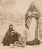 Two individuals dressed in traditional garments, with their faces partially veiled. One person is seated on the ground, appearing to handle a basket containing coiled ropes or similar items, while using a rod-like object. The other individual stands nearby, holding a piece of cloth or shawl. The background features a painted mural depicting a natural landscape with trees.