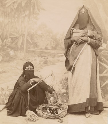Two individuals dressed in traditional garments, with their faces partially veiled. One person is seated on the ground, appearing to handle a basket containing coiled ropes or similar items, while using a rod-like object. The other individual stands nearby, holding a piece of cloth or shawl. The background features a painted mural depicting a natural landscape with trees.