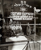 A storefront with the signage reading 'Chicken Market' in both Hebrew and English. A series of plucked chickens hang by their feet in the window display, with a silhouette of a person visible behind the counter. Decorative elements include a rooster and a duck illustrated on the glass.