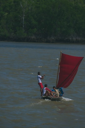 Three individuals are on a small wooden boat with a vibrant red sail, moving across a body of water. The boat is tilted slightly due to the wind filling the sail. The people on the boat are actively engaged in steering and managing the sail. Dense green foliage lines the far bank, indicating a lush, natural environment.