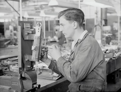 A person in work attire is operating machinery in a workshop setting, focusing intently on the task at hand. The background is filled with various tools and equipment, suggesting a busy, industrial environment.