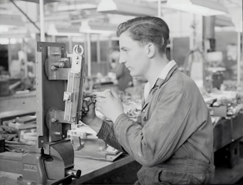 A person in work attire is operating machinery in a workshop setting, focusing intently on the task at hand. The background is filled with various tools and equipment, suggesting a busy, industrial environment.