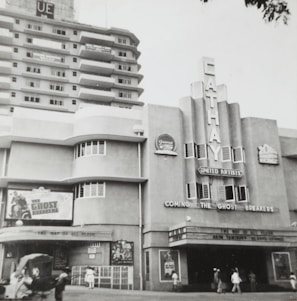 A black and white photograph of an old cinema building labeled 'Cathay' with art deco architectural features. The marquee advertises a movie titled 'The Ghost Breakers'. Pedestrians walk by on the street in front of the cinema.